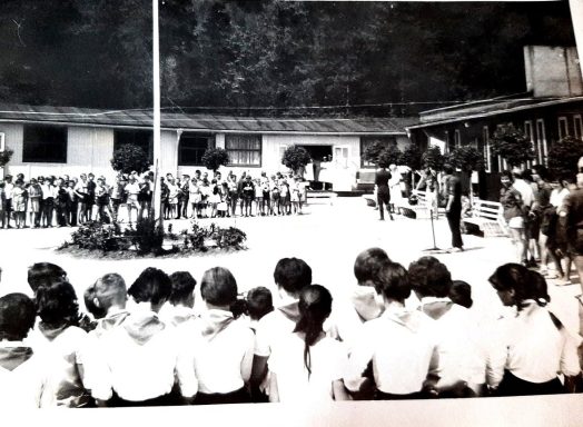 Schüler sitzen vor einer Versammlung im Freien, mit Bäumen und Gebäuden im Hintergrund.