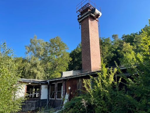 Verfallenes Gebäude mit einem hohen Kamin, umgeben von Bäumen und blauem Himmel.